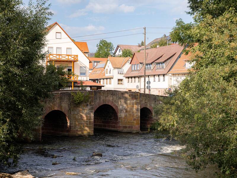 Eine alte Steinbrücke über einem Fluss, im Hintergrund Häuser.
