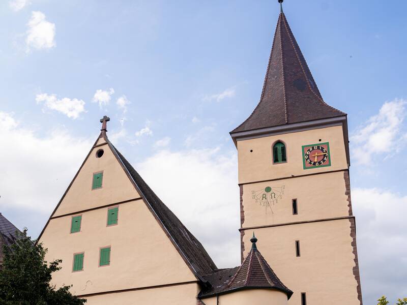 Das Bild zeigt eine historische Kirche mit einem hohen Turm, einem Uhrzifferblatt und umgeben von Bäumen, vor einem leicht bewölkten Himmel.